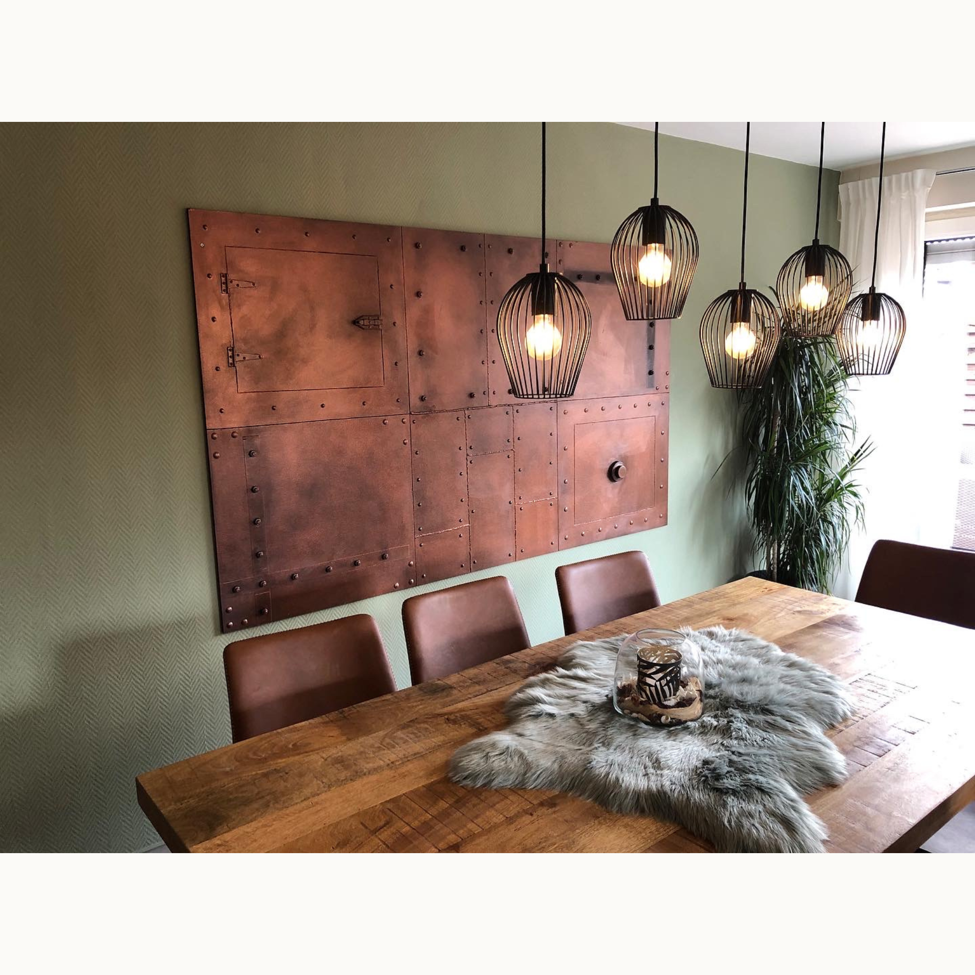 Esszimmer mit großem Kupfer-Wandpaneel mit Nieten über einem Holztisch mit Käfig-Hängeleuchten
